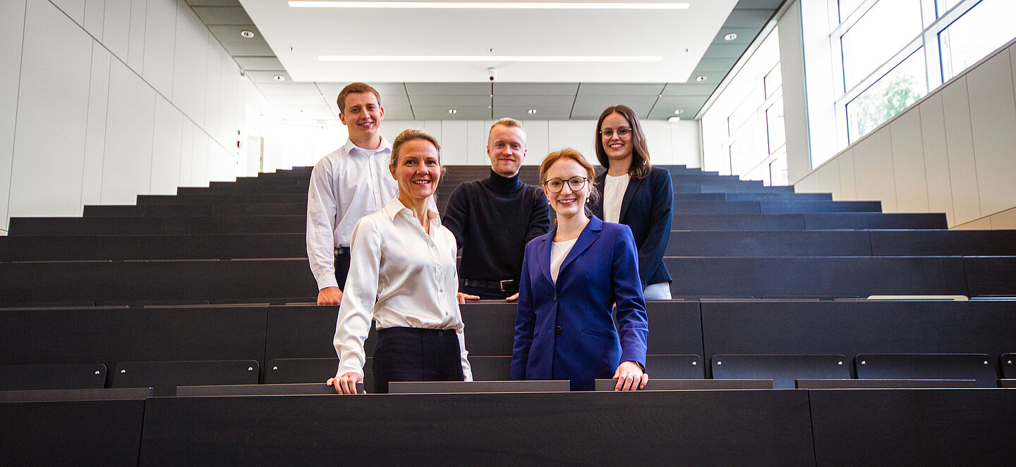 Das Projektteam in einem Hörsaal der HTWK, aufgestellt in zwei Sitzreihen. Hintere Reihe, von links nach rechts: Herr Leon Lowak, Herr Max Neumann und Frau Sophie Gehringer. Vordere Reihe, von links nach rechts: Fr. Prof. Dr. Manger-Nestler und Frau Clara Schneidenbach.